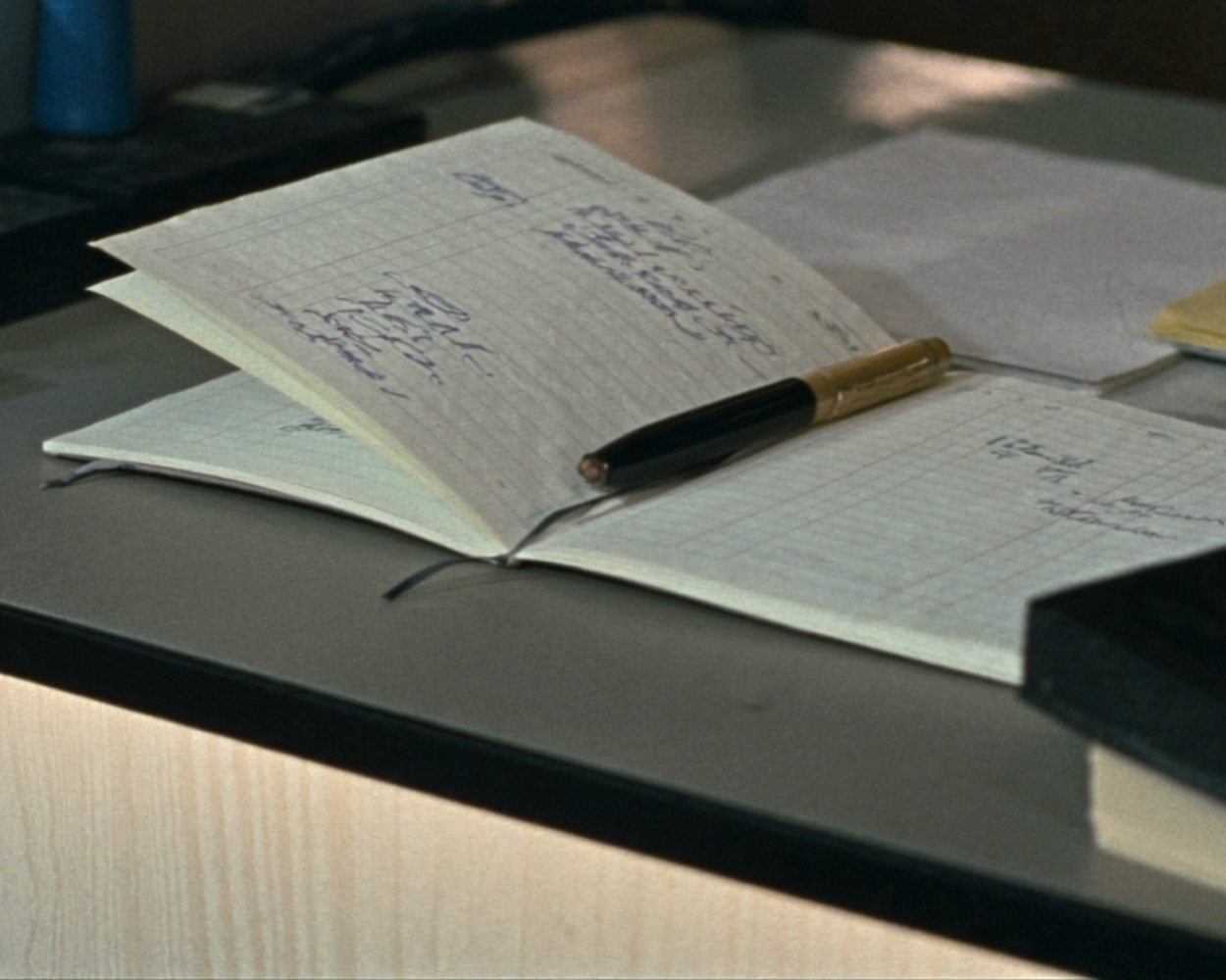 A worn paper appointment book open on a front-desk counter beside a pen, a rubber stamp and a short stack of file folders, the handwriting too soft to read.