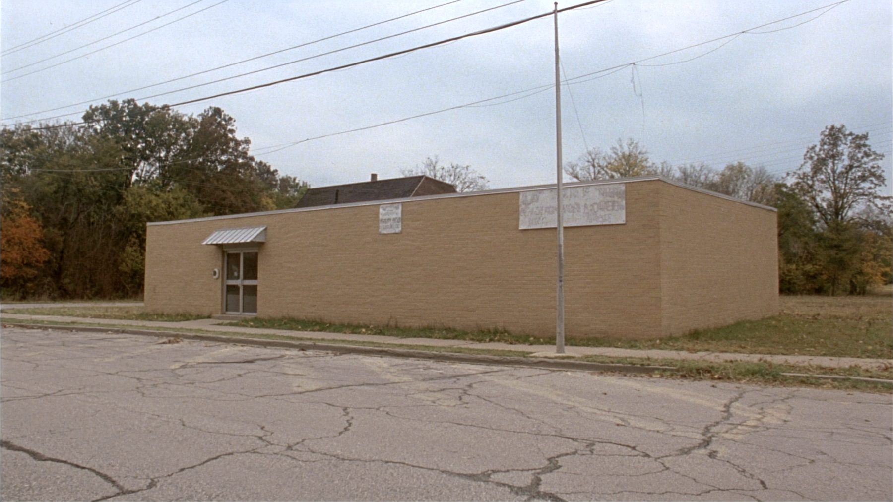 A small single-storey brick medical office on a quiet small-town street under flat autumn daylight, with two parking spaces and a bare sign board.
