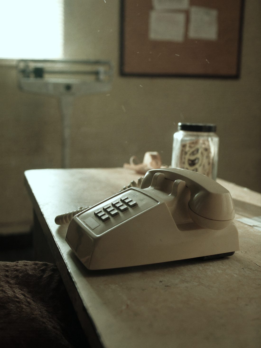 A corded telephone on a laminate counter beside a jar of tongue depressors and a small floor scale in an old medical office.