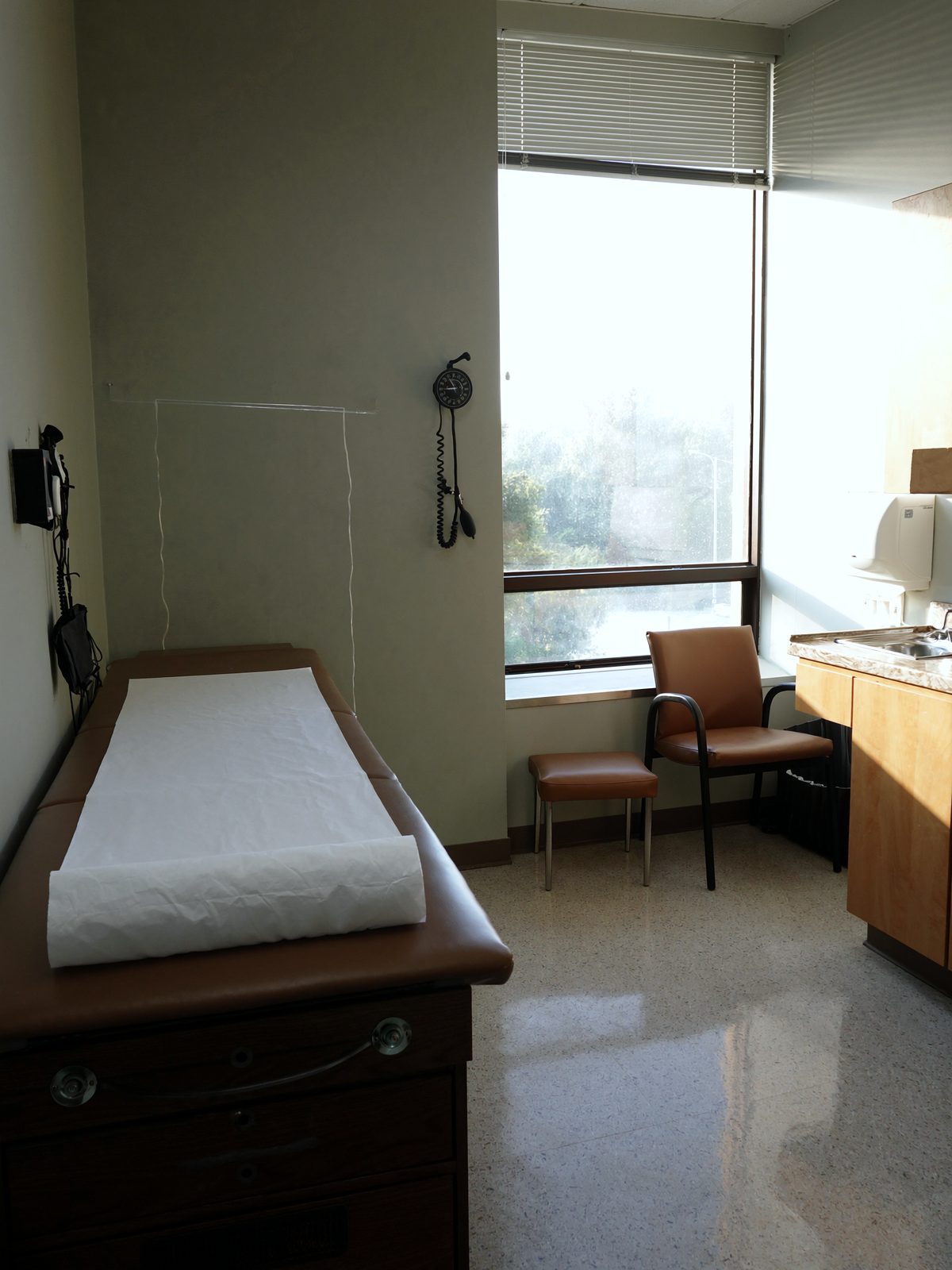 A corner of a small exam room in the late morning: paper-covered table, a low stool, and a manual blood pressure cuff on the wall.